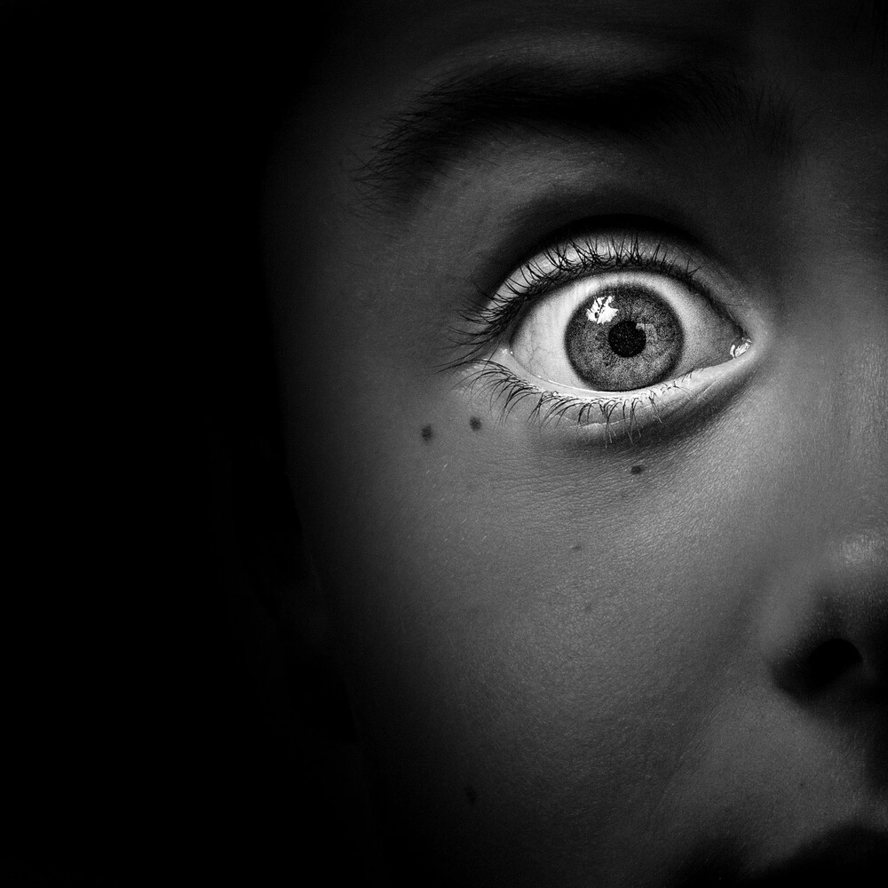 Black and white close-up of a wide, startled eye with a visible catchlight, half of the face lost in deep shadow.