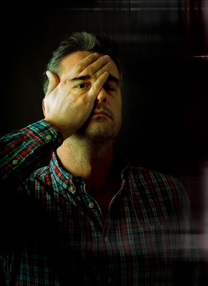 Man in a plaid shirt pressing his hand over his eyes against a dark background, lit by dramatic low-key light.