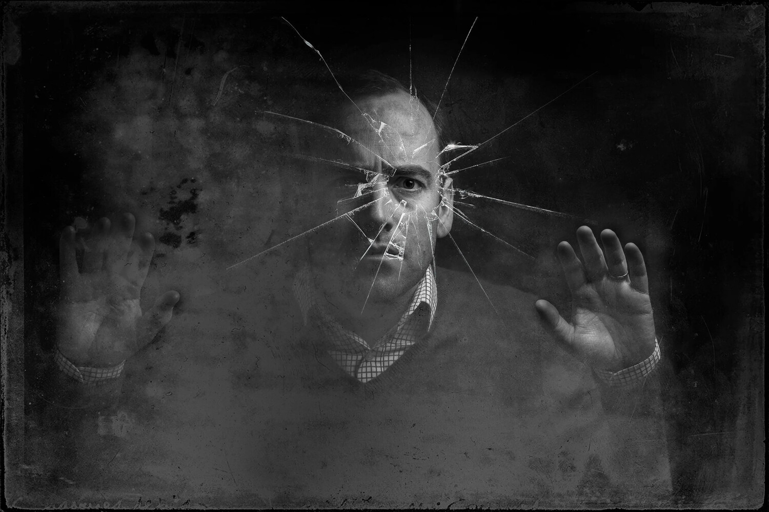 Black and white portrait of a man behind a cracked glass pane, with radiating fractures across his face and his palms pressed against the glass.