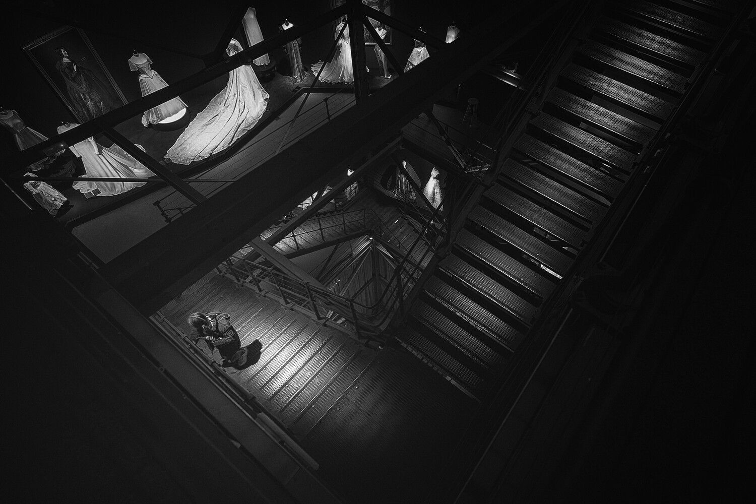 Black and white high-angle view down a metal staircase toward a lit landing, with white gowns on mannequins visible behind the railing in the upper left.