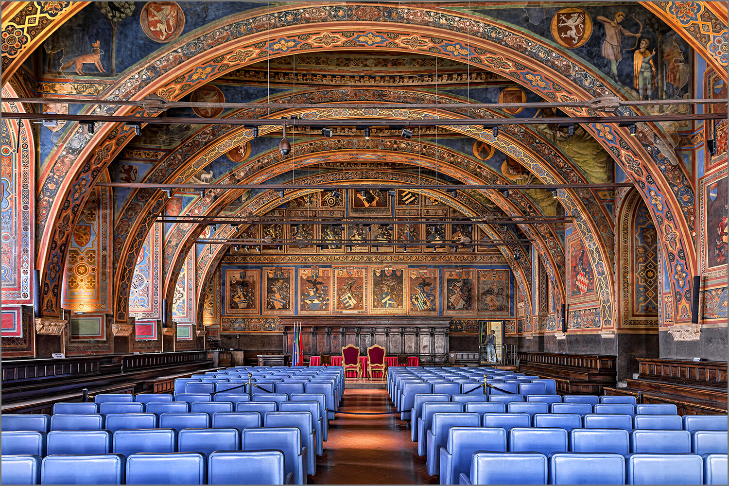 Ornate chapel interior with concentric barrel vaults decorated in gold and blue, rows of blue seats facing the altar