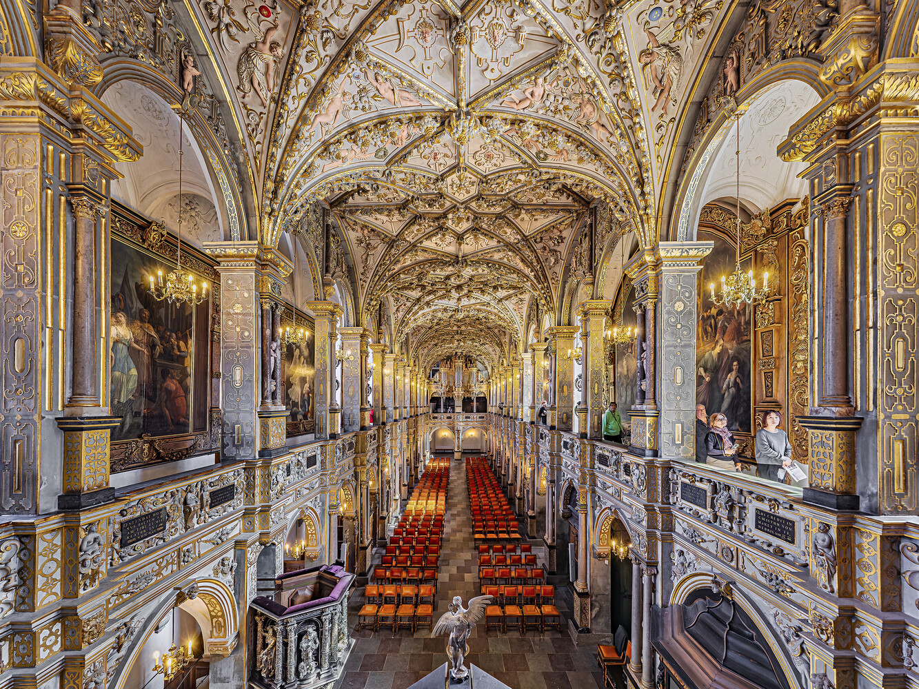 Ornate historic hall with gilded balconies, a painted vaulted ceiling, and rows of red chairs leading toward a white marble statue.
