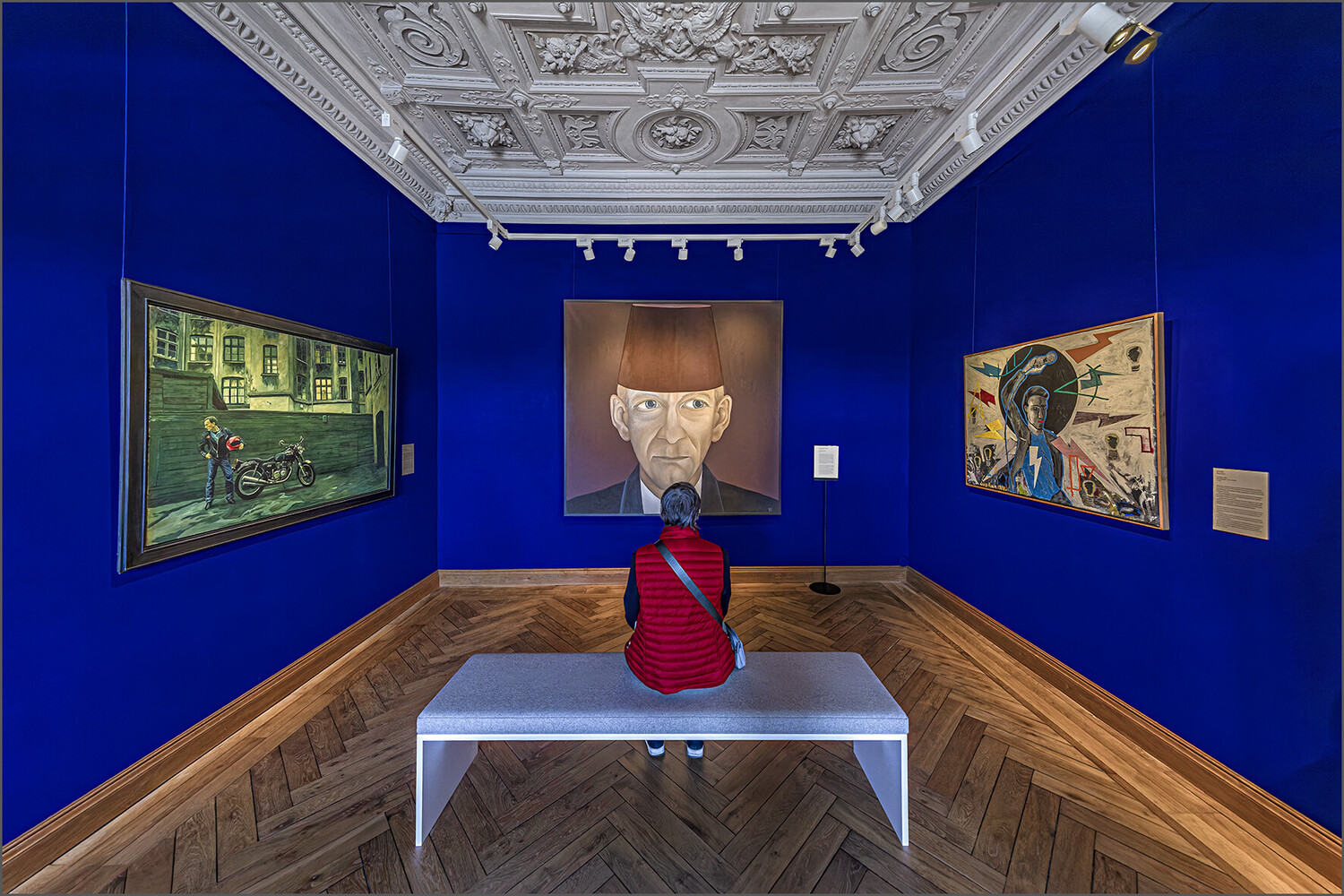 Visitor in a red jacket seated on a bench facing a central portrait of an older man in a gallery with deep blue walls