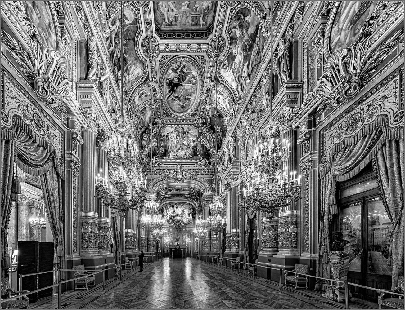Black and white view down an ornate gilded hall with crystal chandeliers, painted ceilings, and a long parquet floor