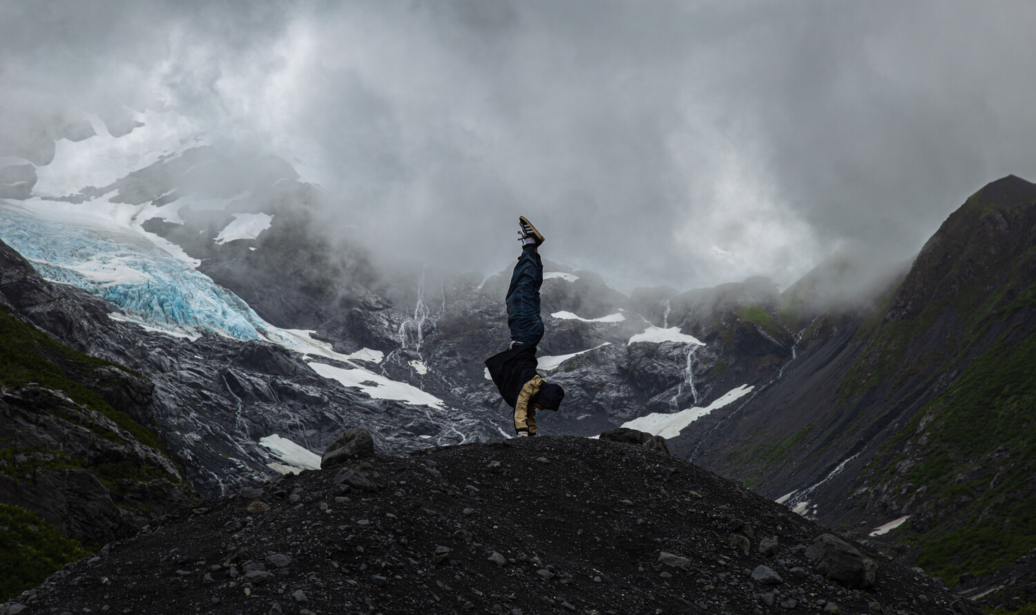 Lone hiker standing on dark rocky terrain facing a glacier and misty mountain valley