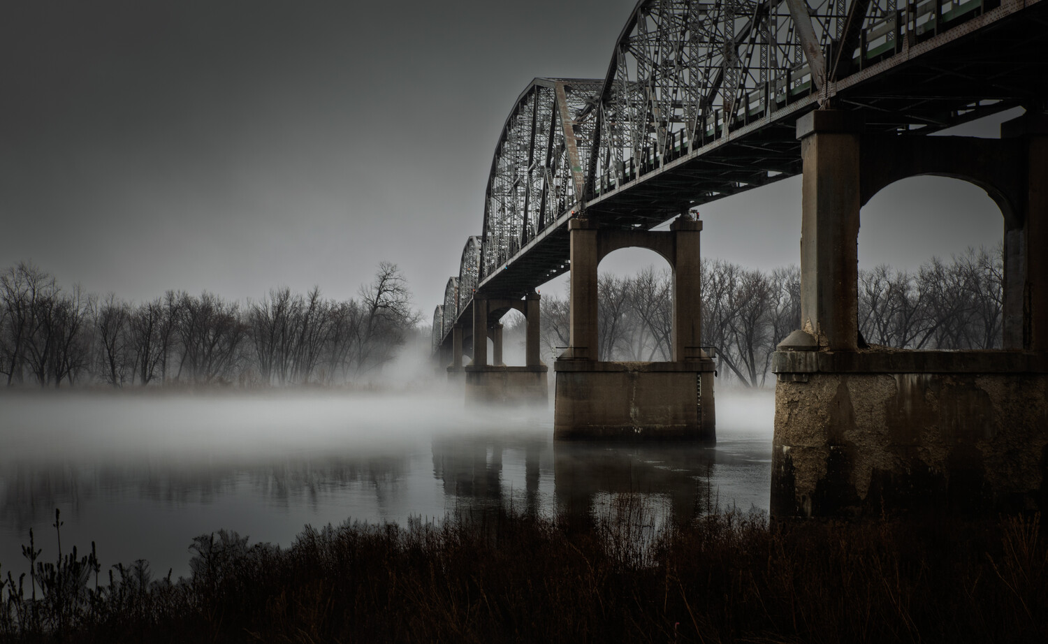 Black and white view of a steel truss bridge arching over a fog-covered river, with bare trees along the bank.