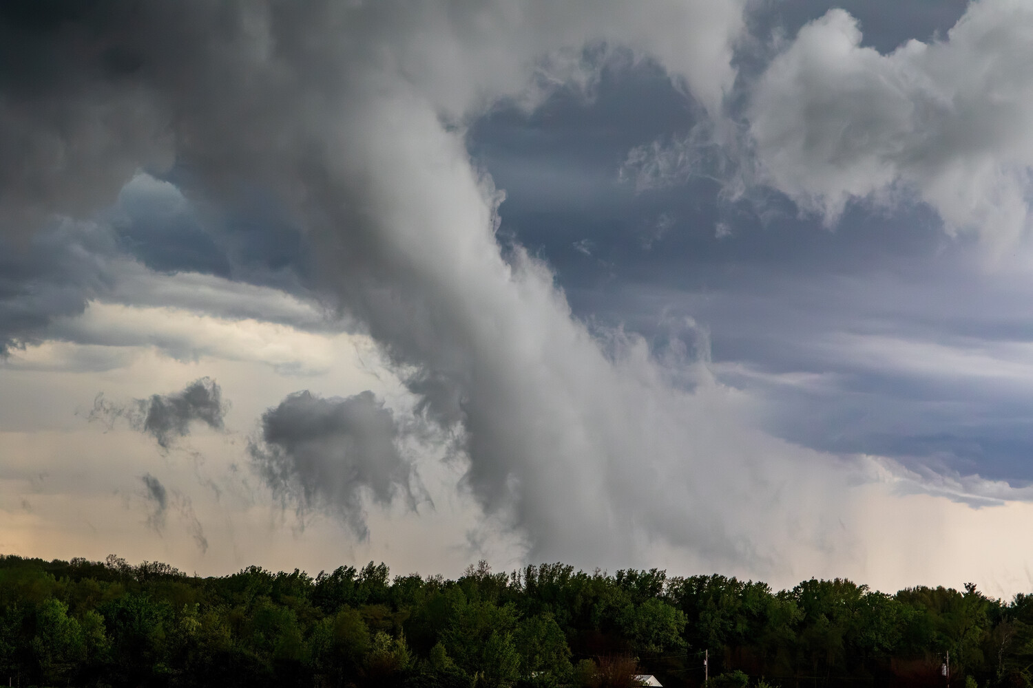 Dramatic funnel-like storm cloud descending from a dark, overcast sky above a dense green tree line