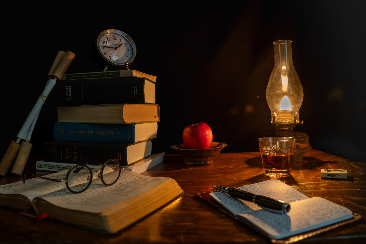Arranged still life with stacked books, vintage clock, oil lamp, apple, open notebooks, telescope, and glass of whiskey on wooden table