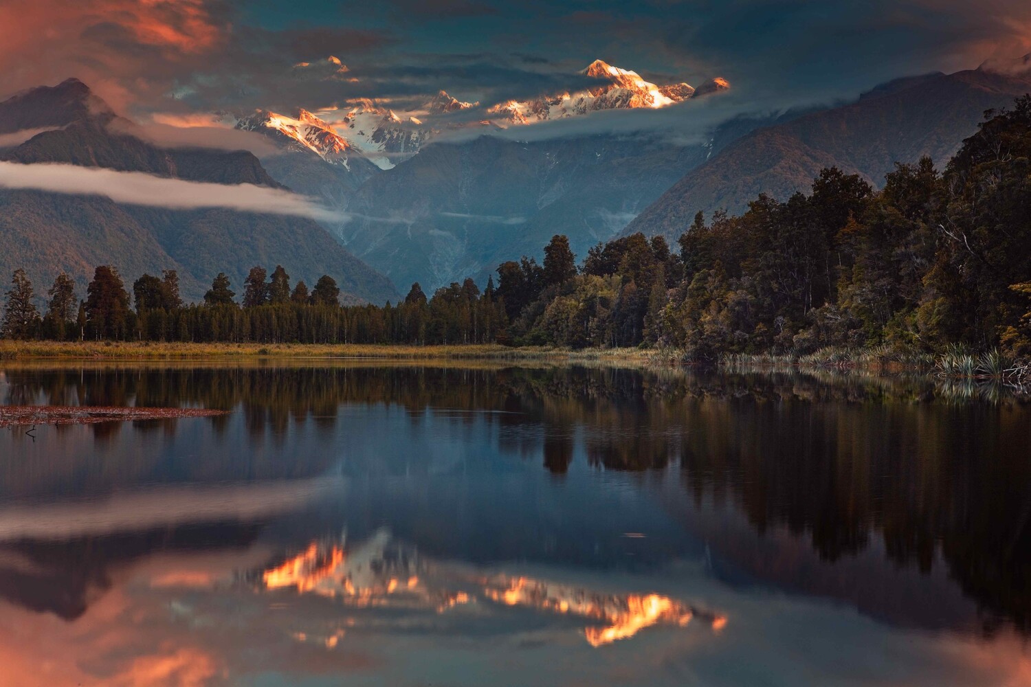Mountain lake at sunset with forested shoreline and snow-capped peaks reflected in still water