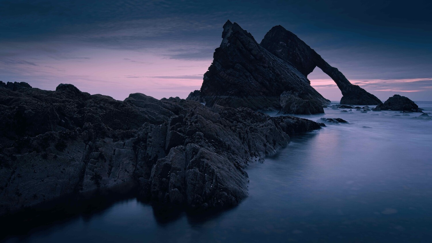Dramatic rock formation rising from smooth water at twilight with purple-pink sky