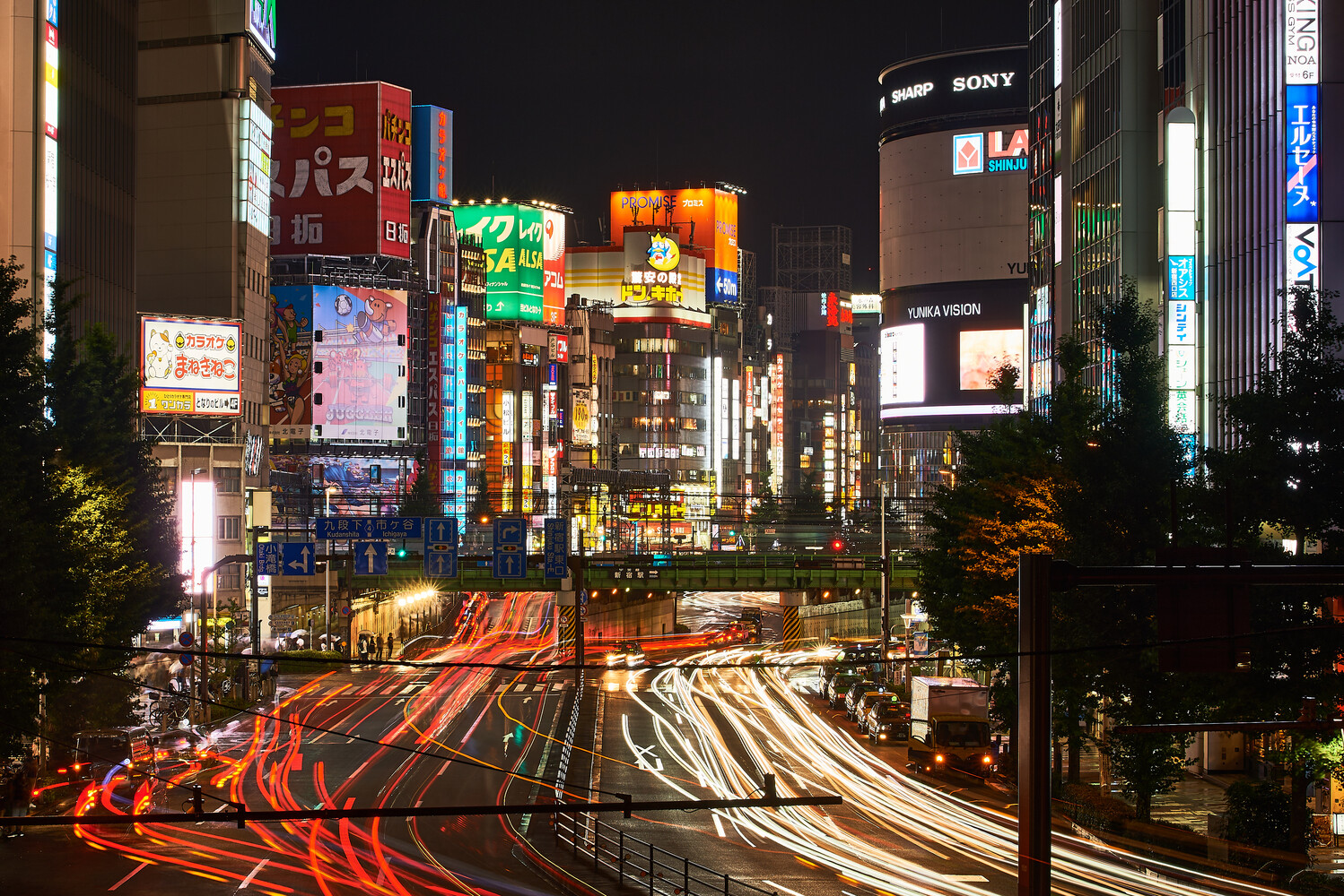 Shibuya Crossing at night with light trails from vehicles streaking across the intersection