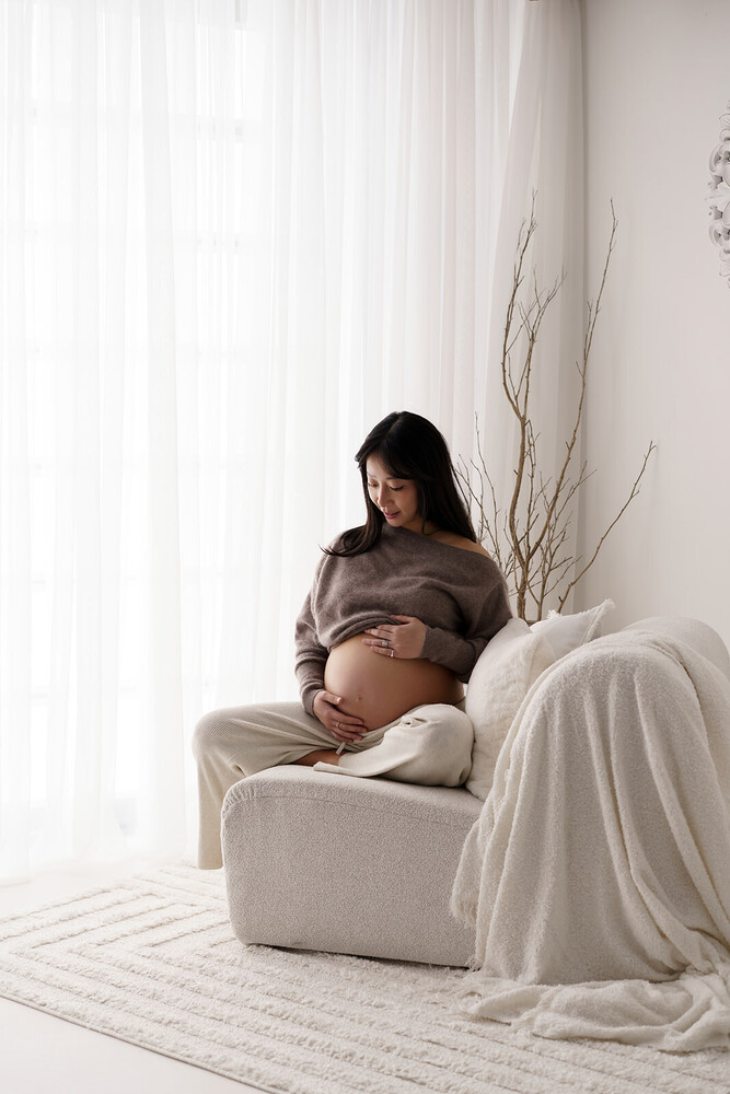 Pregnant woman in neutral tones cradling her belly while seated in a cream armchair