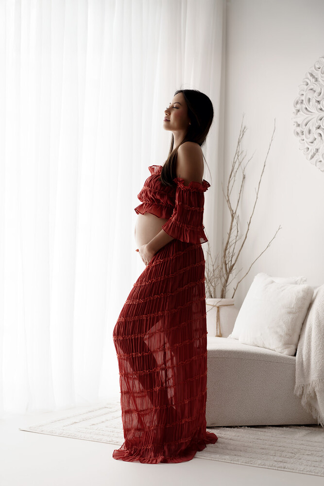 Woman in burgundy textured dress standing by white bed near window with sheer curtains