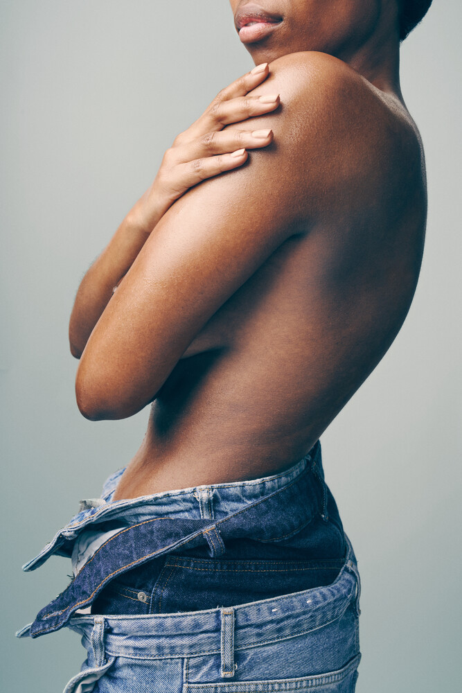 Shirtless person from behind wearing low-rise denim jeans, hand on shoulder against neutral background