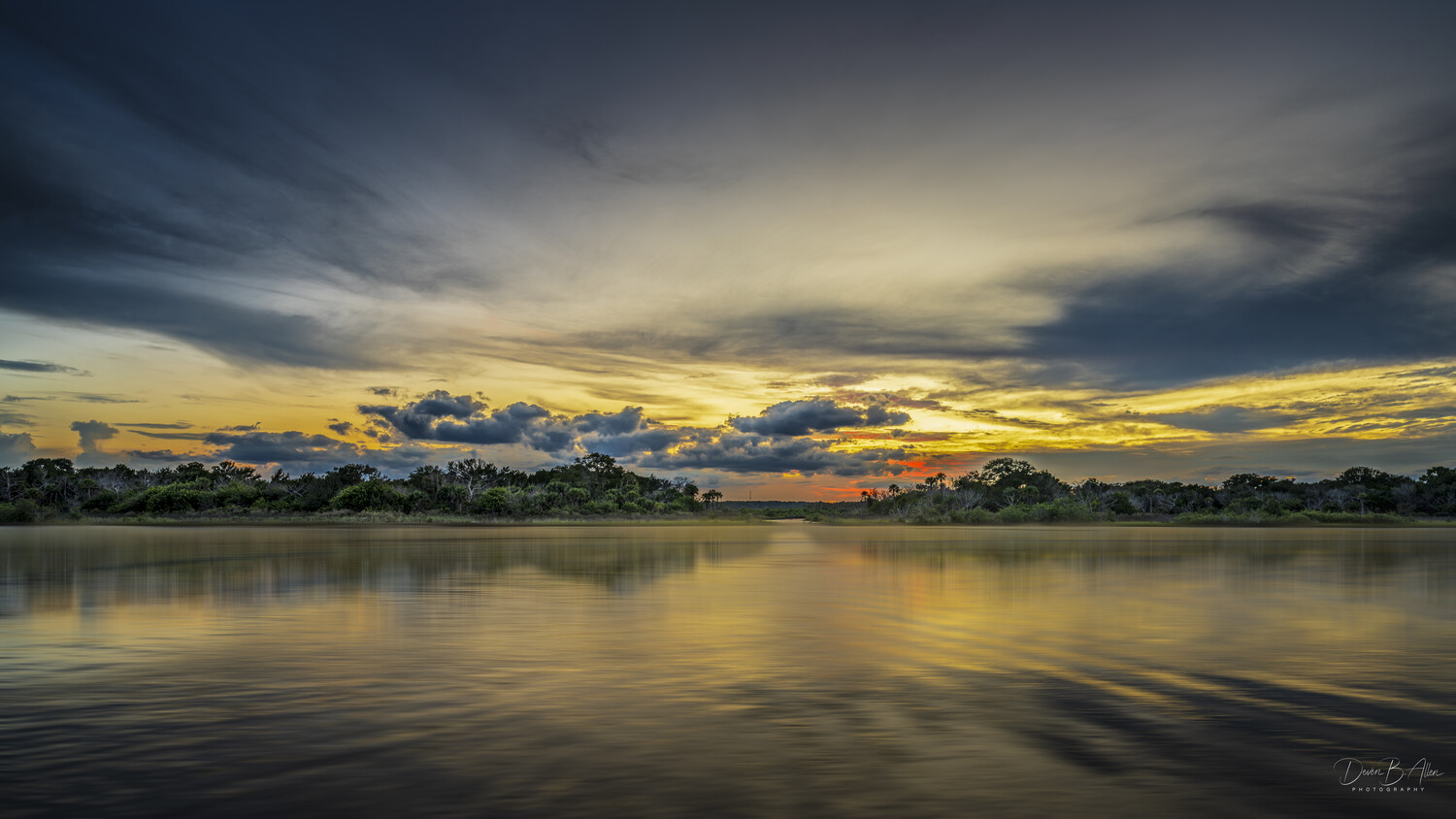 Sunset over a calm river with golden light reflecting off the water and dark clouds above a silhouetted tree line.
