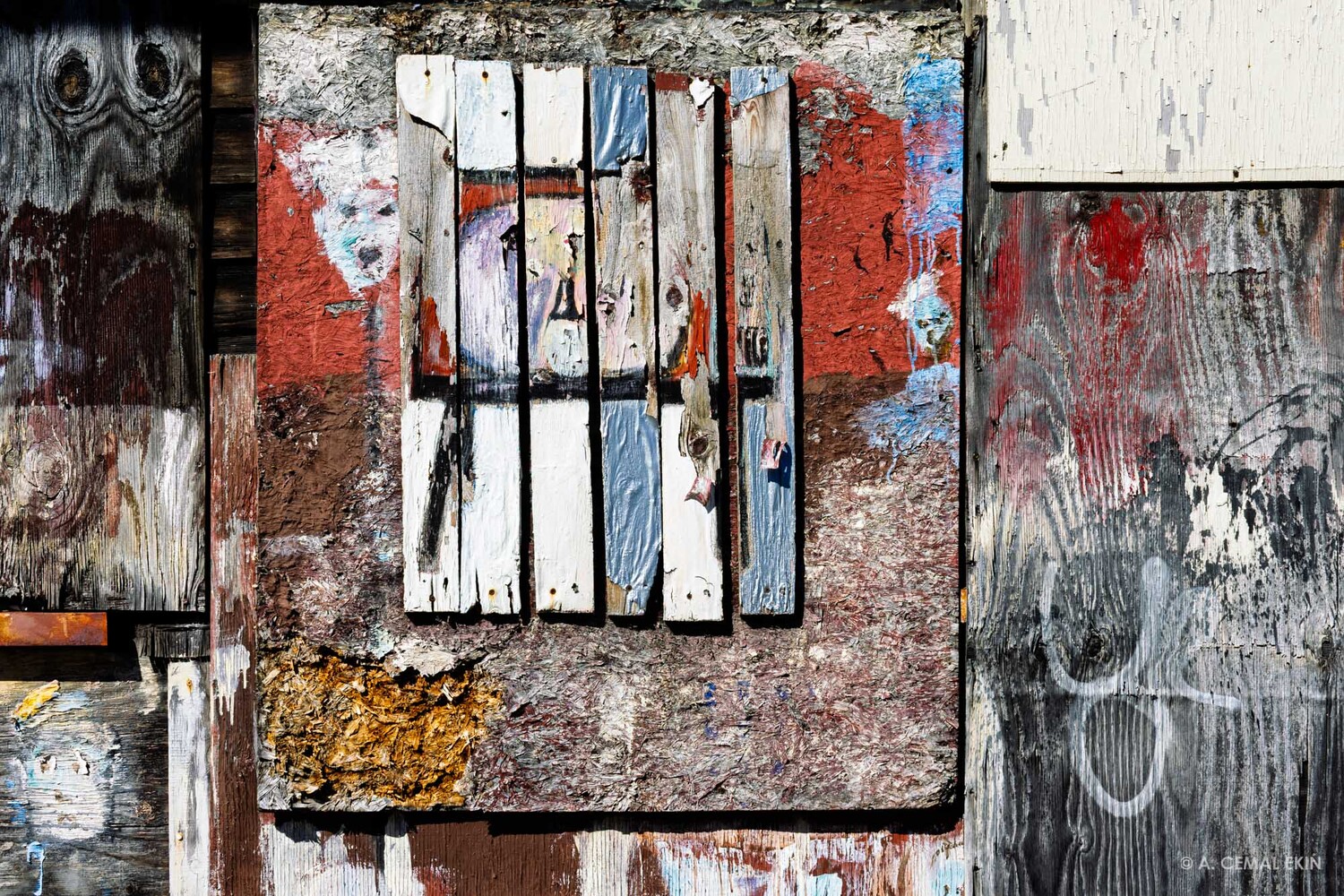 Weathered wooden slats painted white and blue over a red, peeling panel, framed by aged boards with flaking paint and faded graffiti marks.