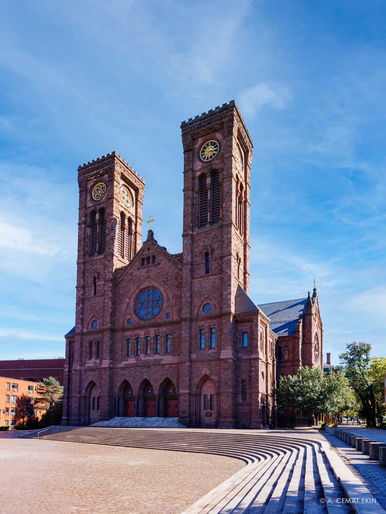 Red brick cathedral with two clock towers and a round stained-glass window under a clear blue sky, viewed from a stone plaza.