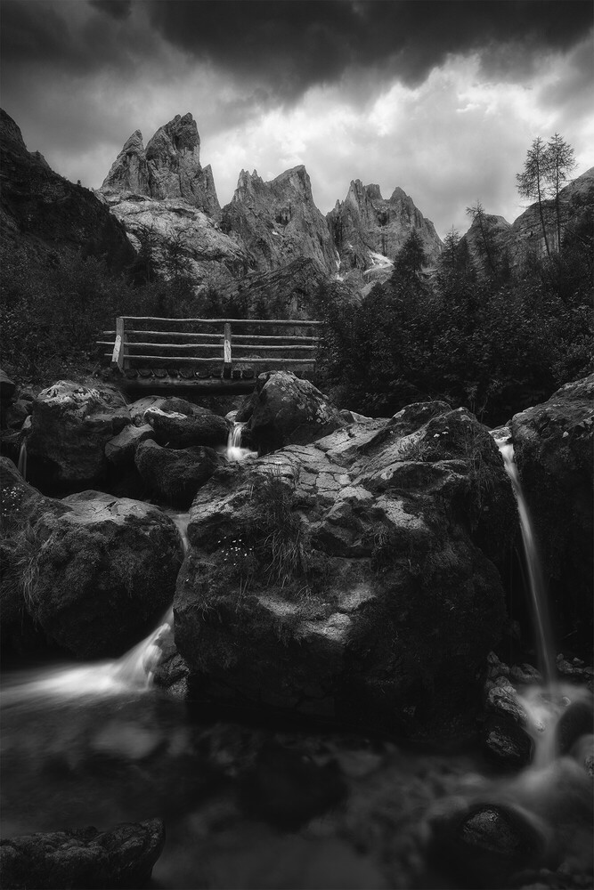 Wooden footbridge spanning a rocky stream beneath dramatic jagged peaks and storm clouds