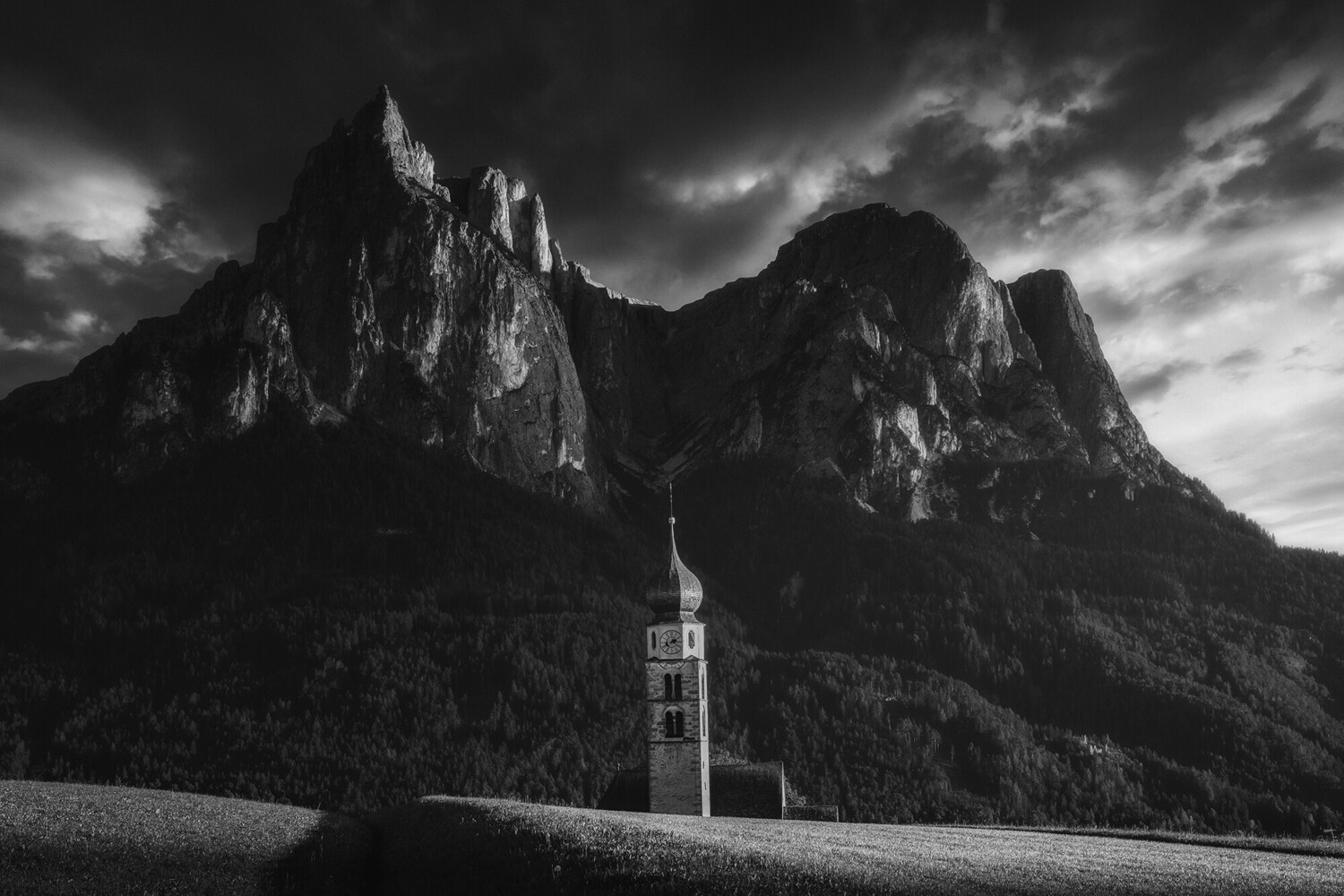 Church with tall steeple nestled in valley beneath dramatic rocky peaks under stormy sky