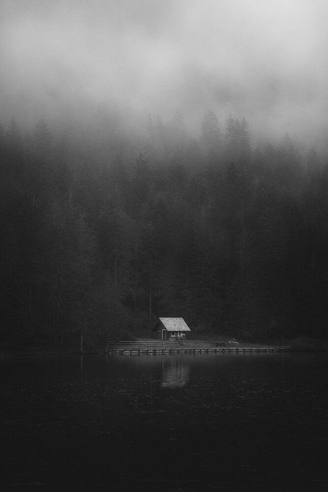 Small wooden cabin with a pitched roof on a jetty above a dark, misty lake, surrounded by fog-shrouded evergreen forest.