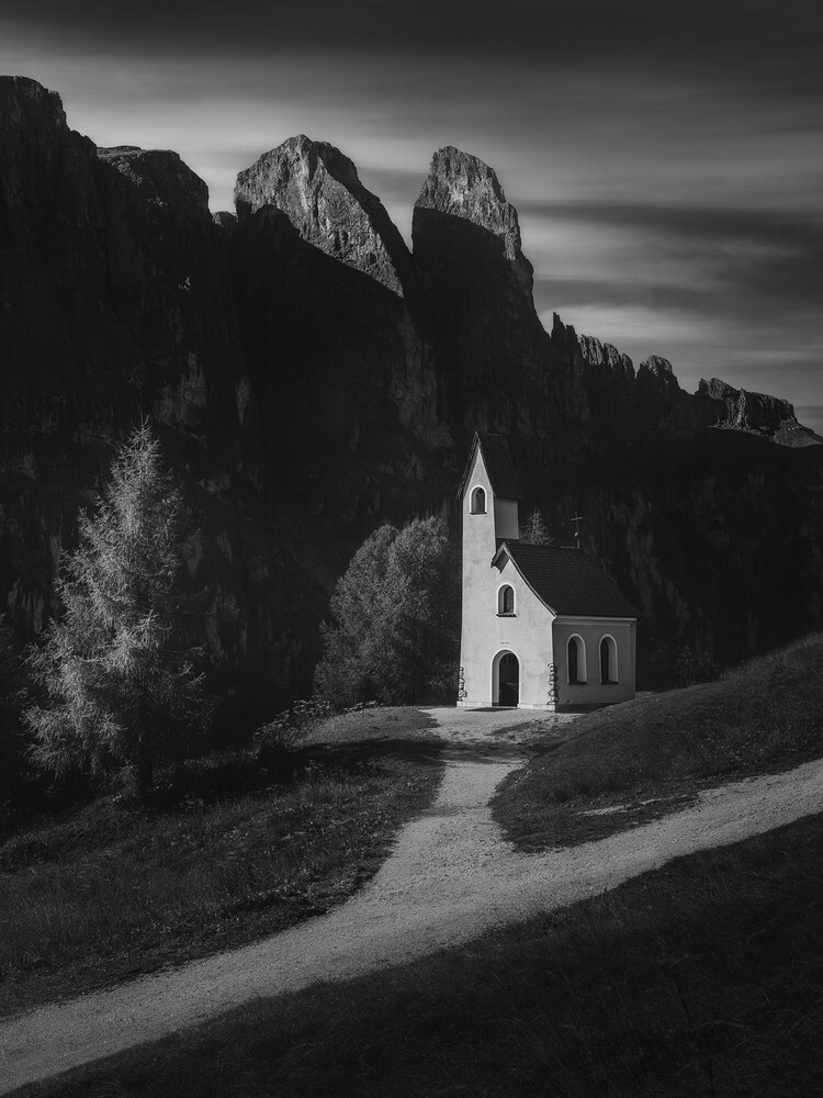 Small white chapel with a pointed roof standing beneath dramatic dark mountain peaks, reached by a winding gravel path, in black and white.