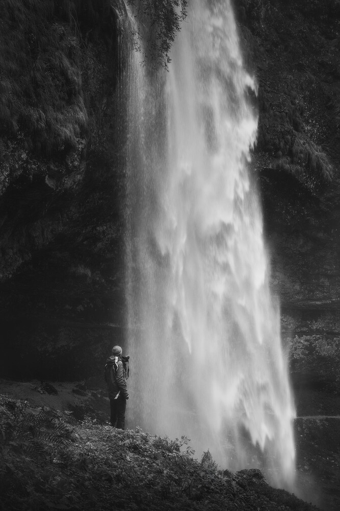 A lone person in a hooded jacket stands at the base of a tall, wide waterfall pouring down a dark cliff, in black and white.