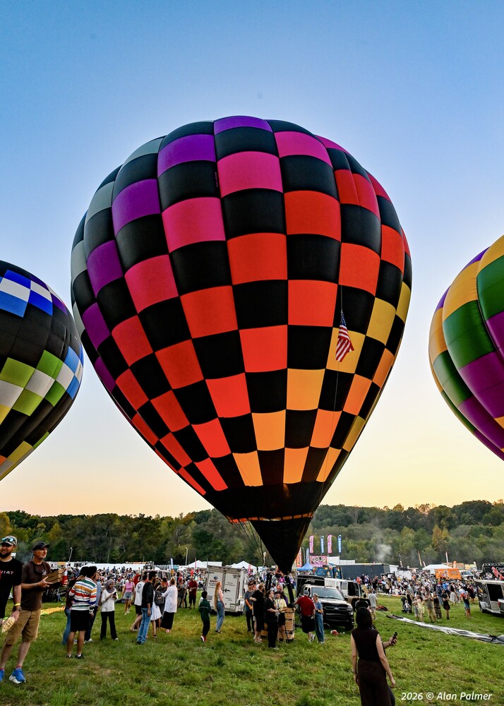 Checkered hot air balloon in black, pink, orange, and purple panels glowing at dusk as a crowd gathers on a grassy field