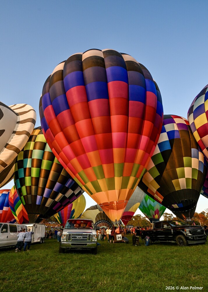 Colorful hot air balloons inflating at dusk on a grassy field, with a large multicolored balloon in the center and trucks parked nearby.