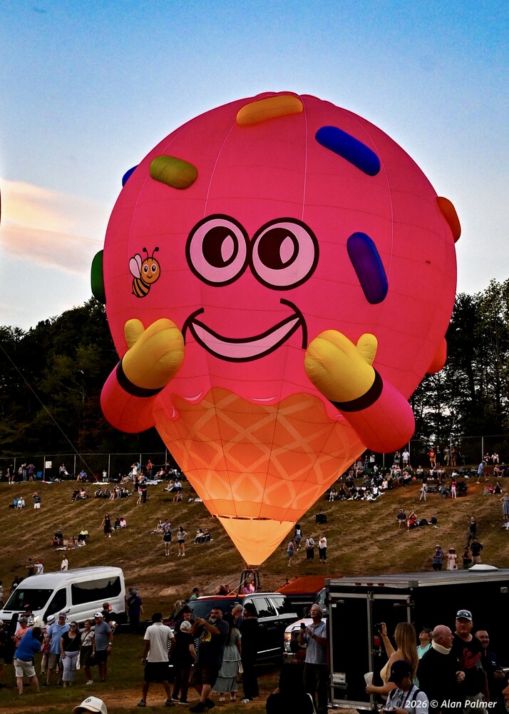 Pink ice cream cone-shaped hot air balloon with sprinkles and a smiling face, glowing at dusk above a crowd on a grassy field