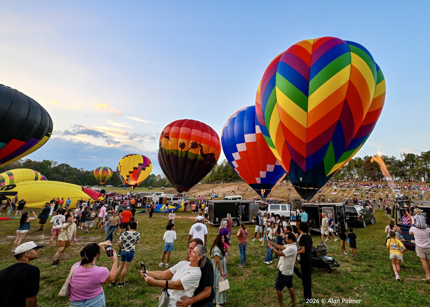 Crowd watches colorful hot air balloons inflating on grass at dusk, with a rainbow chevron balloon in the foreground right.