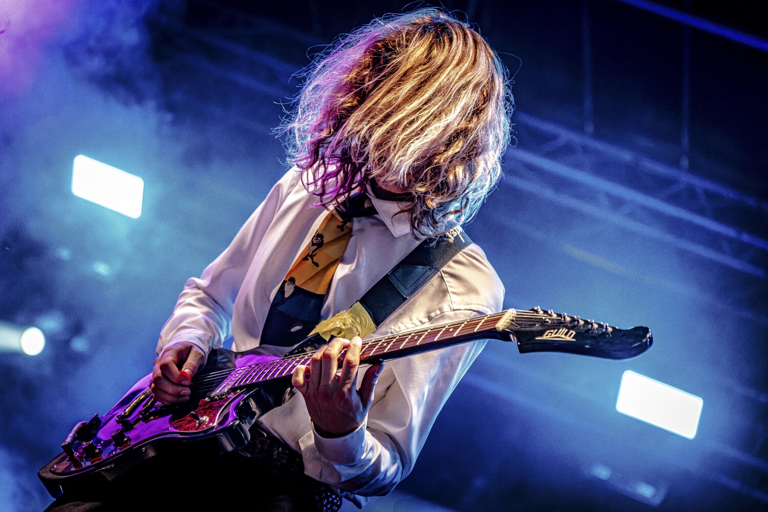 Musician with long blonde hair playing electric guitar on stage under blue and purple stage lighting