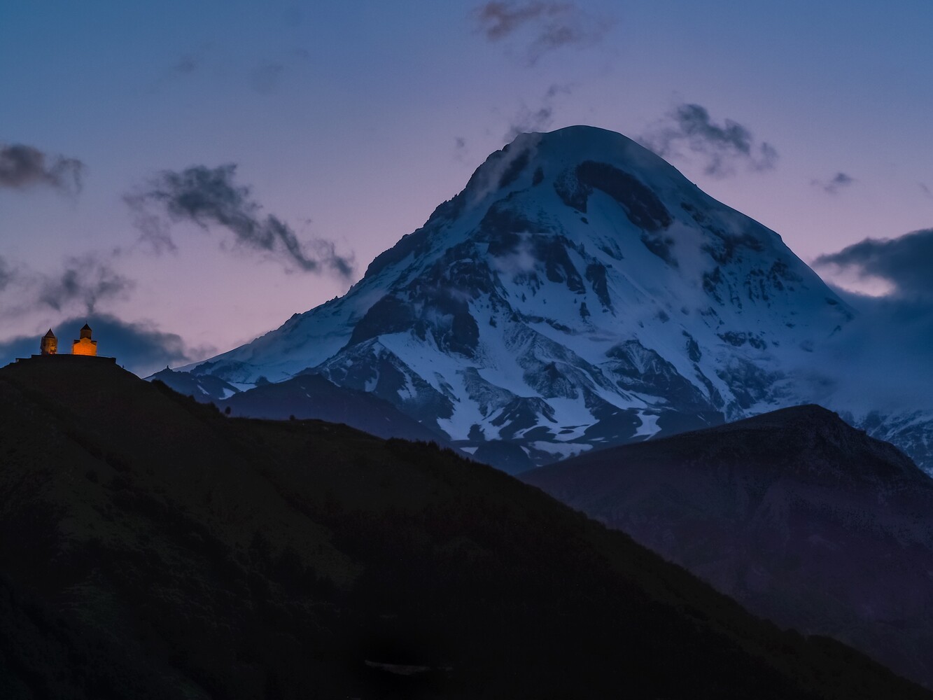 Snow-covered volcanic mountain at twilight with warm-lit structure on distant ridge