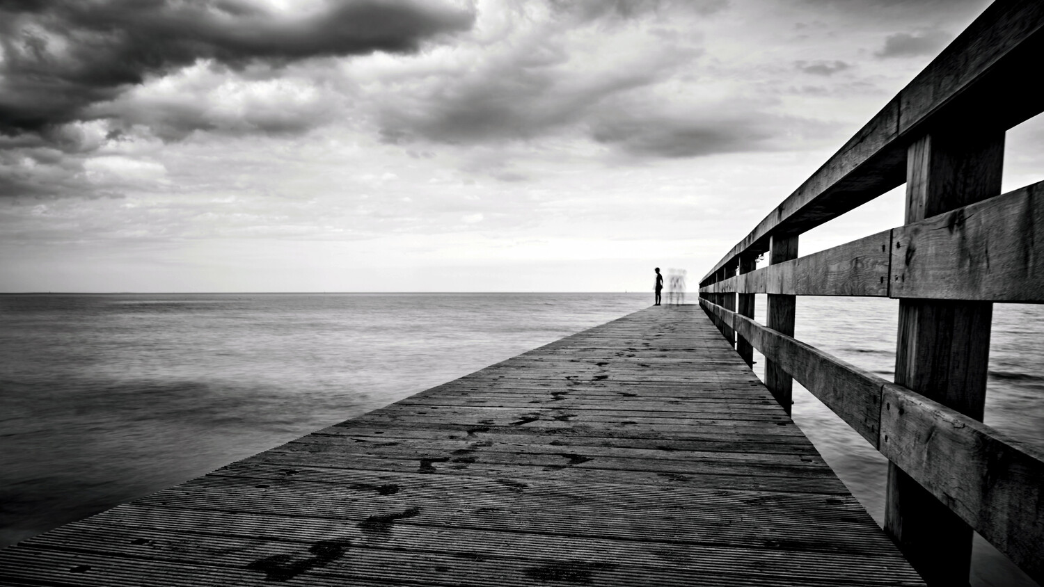 Wooden pier extending into calm ocean under dramatic cloudy sky with solitary figure in distance