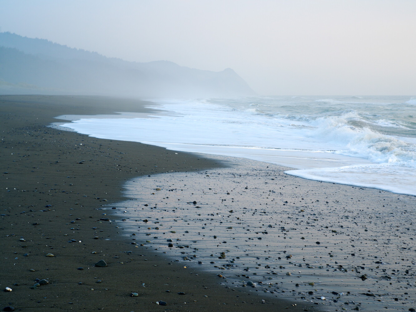 Misty coastal landscape with dark volcanic sand beach and rolling ocean waves