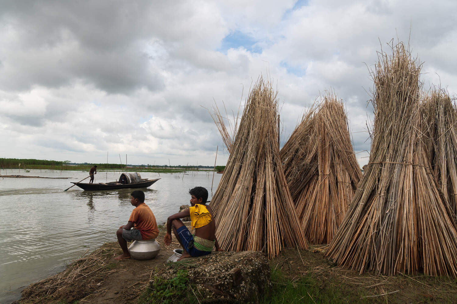 Two people crouching on muddy ground beside bundled reeds, with a boat and water visible in the background