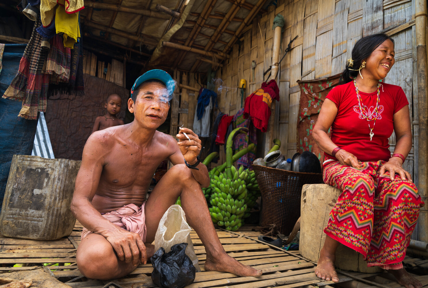Man in blue cap and woman in red shirt sitting inside a modest wooden dwelling