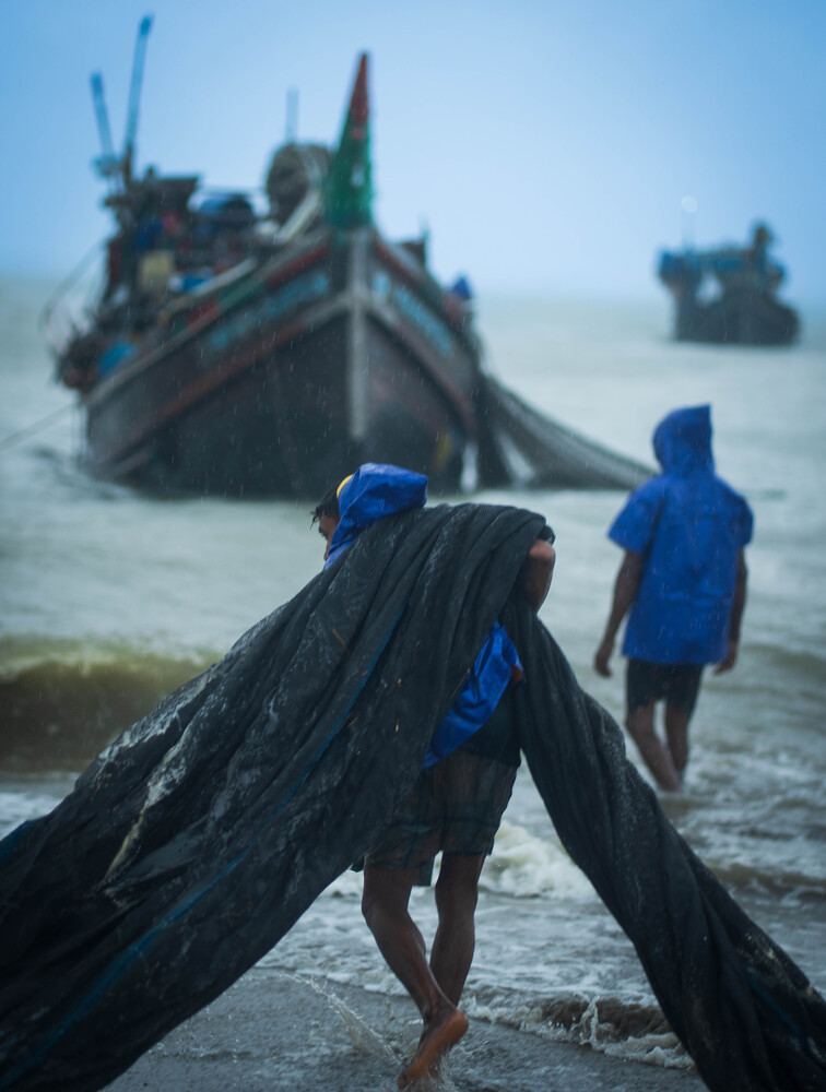 Two children in blue clothing carrying fishing nets toward wooden boats in shallow coastal water