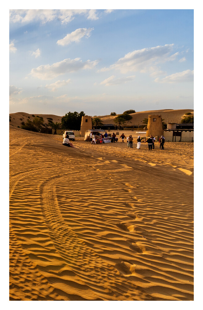 Tourist caravan traveling across rippled golden sand dunes beneath a blue sky with scattered clouds