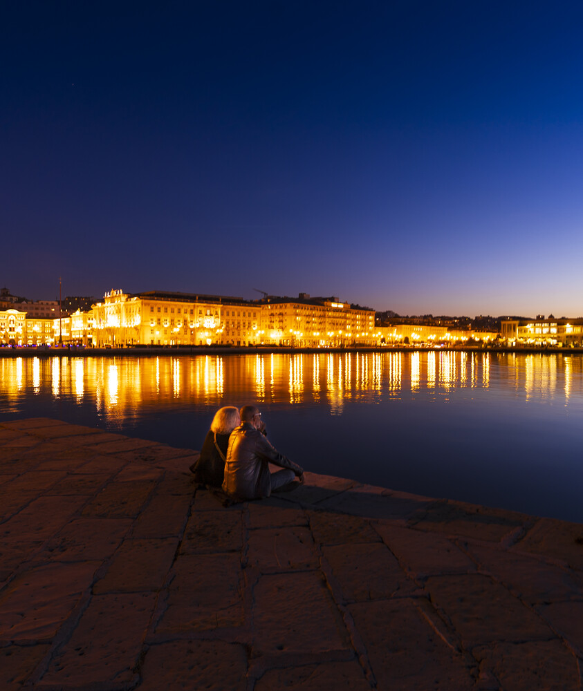 Solitary figure sitting on wet pavement beside illuminated waterfront buildings at dusk