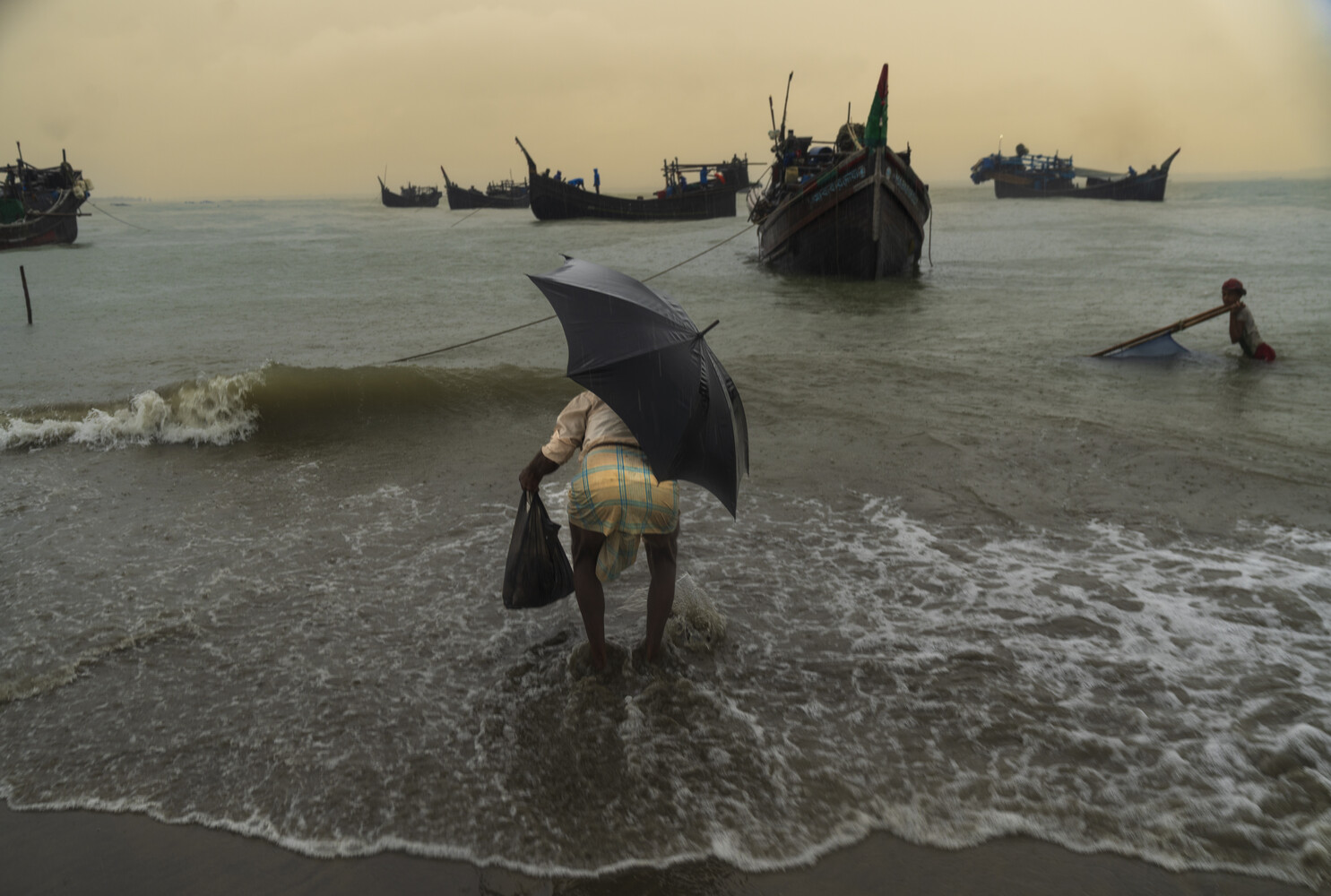 Man with umbrella wading in shallow water with traditional fishing boats anchored offshore