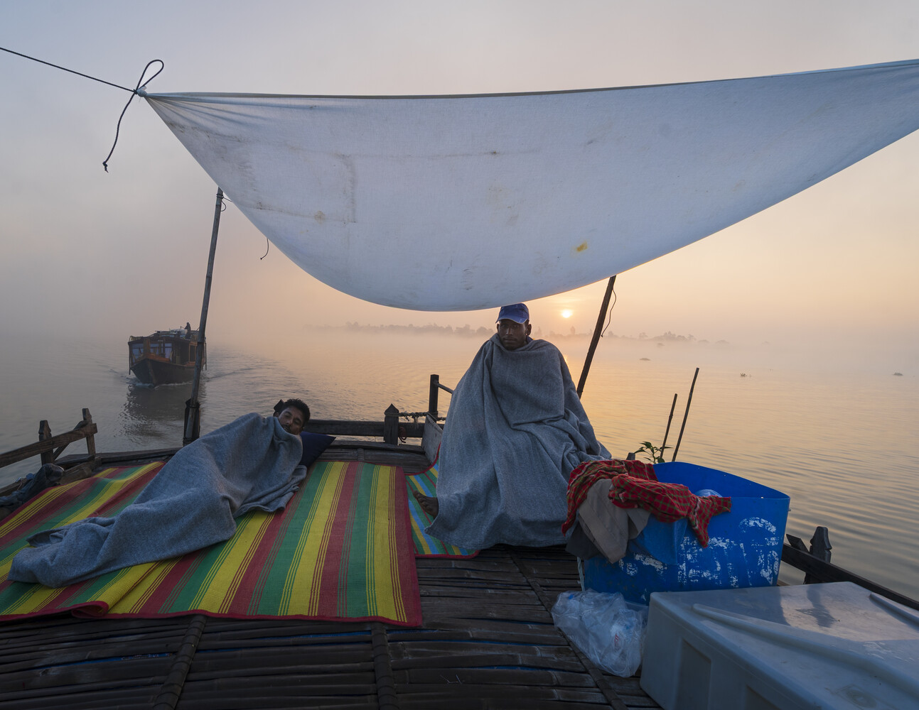 Fisherman in blue wrap sitting on wooden boat at dawn with draped sail and colorful striped fabric