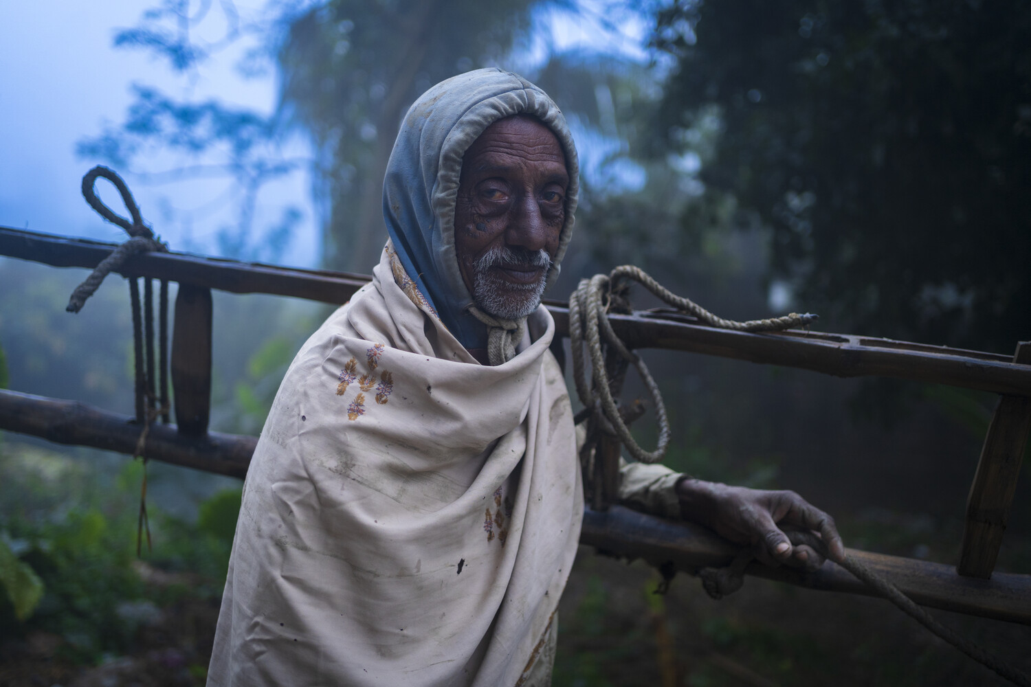 Elderly bearded man in a grey head wrap and white shawl leaning on a rope-bound wooden railing in misty woods