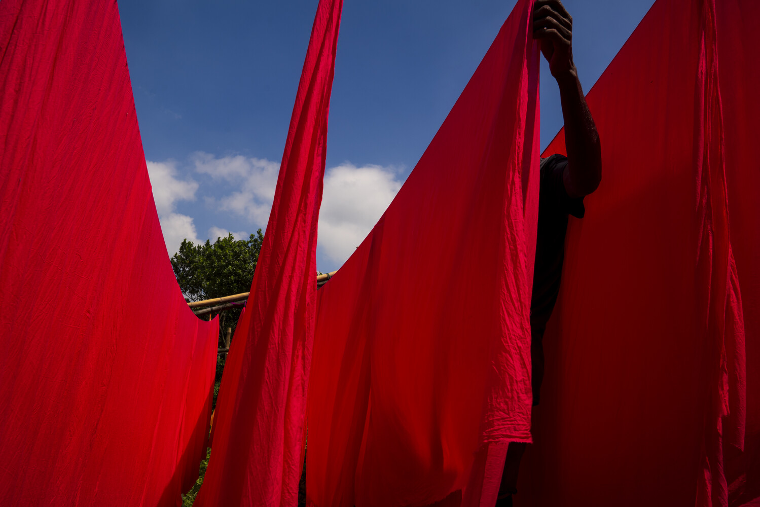 Tall sheets of bright red fabric hanging vertically against a blue sky, with a person's raised arm holding one sheet at the top right.