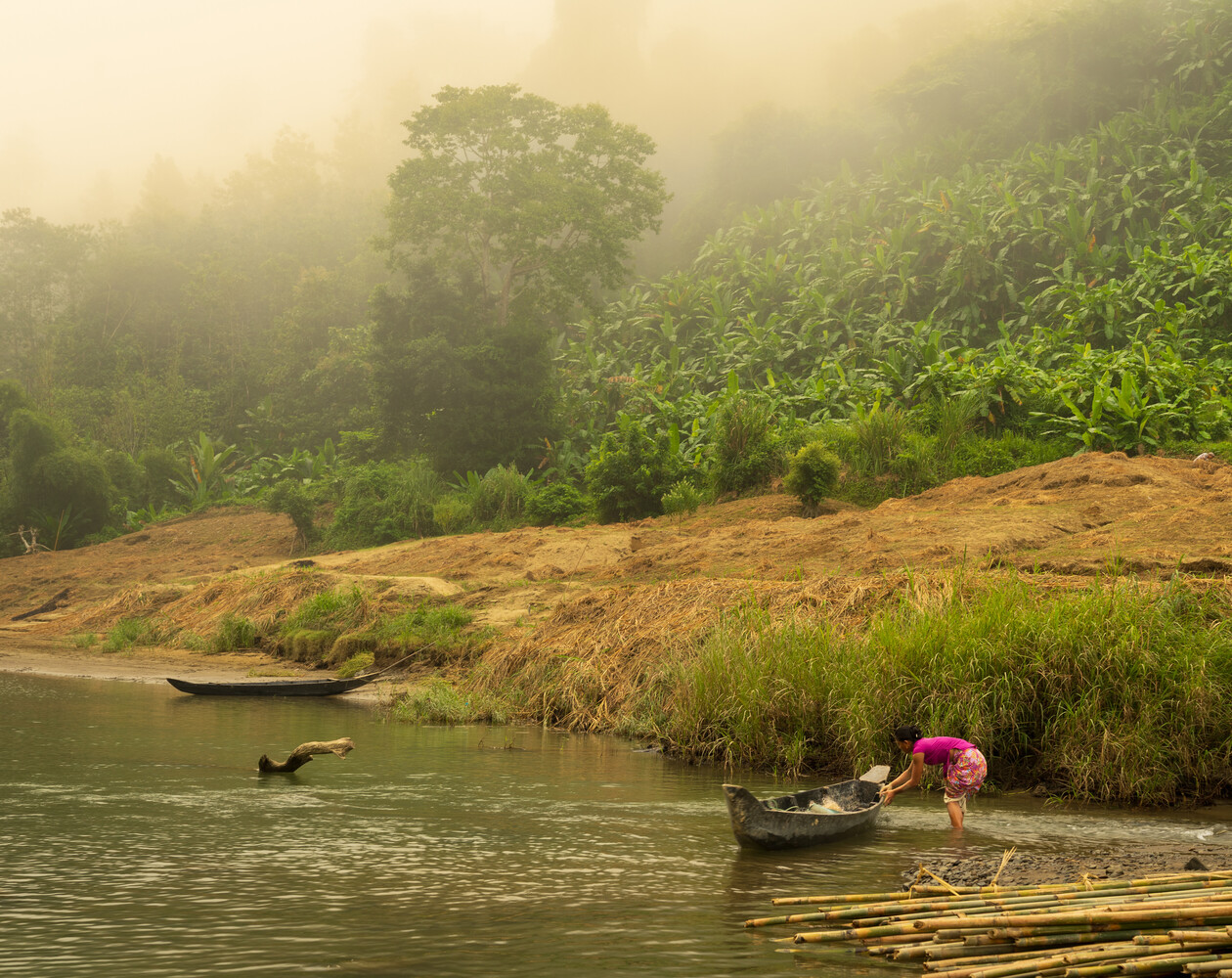 Person in pink clothing pulling a wooden canoe onto a misty riverbank beneath a steep green hillside of banana plants and trees