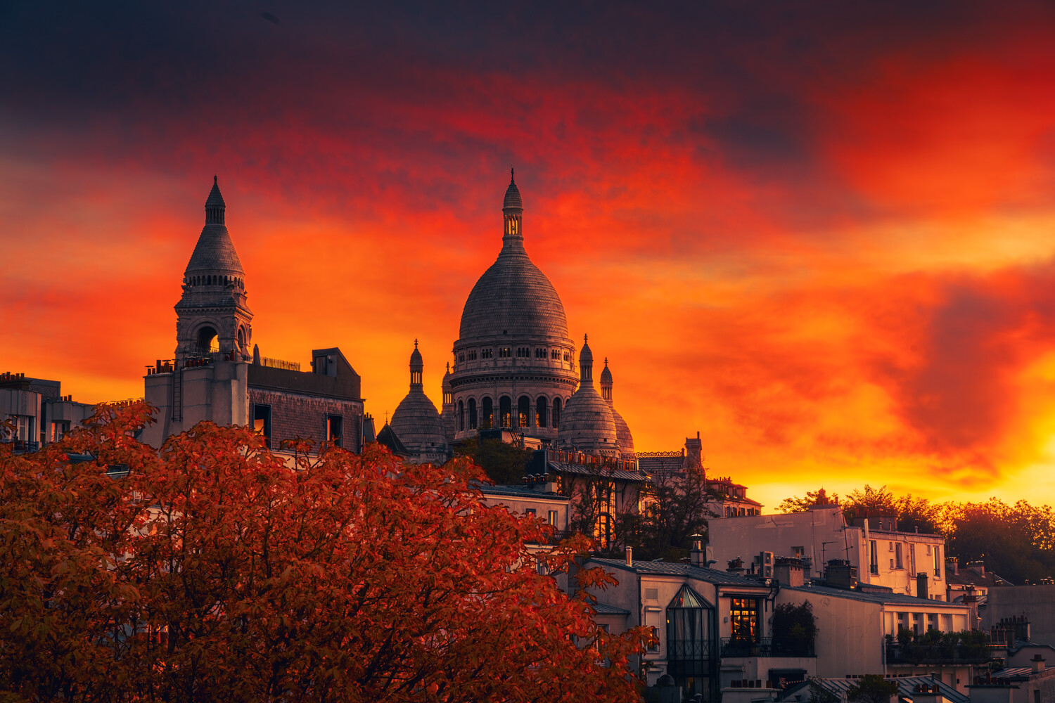 Sacré-Cœur basilica domes rise against a fiery red and orange sunset sky, with autumn foliage in the foreground