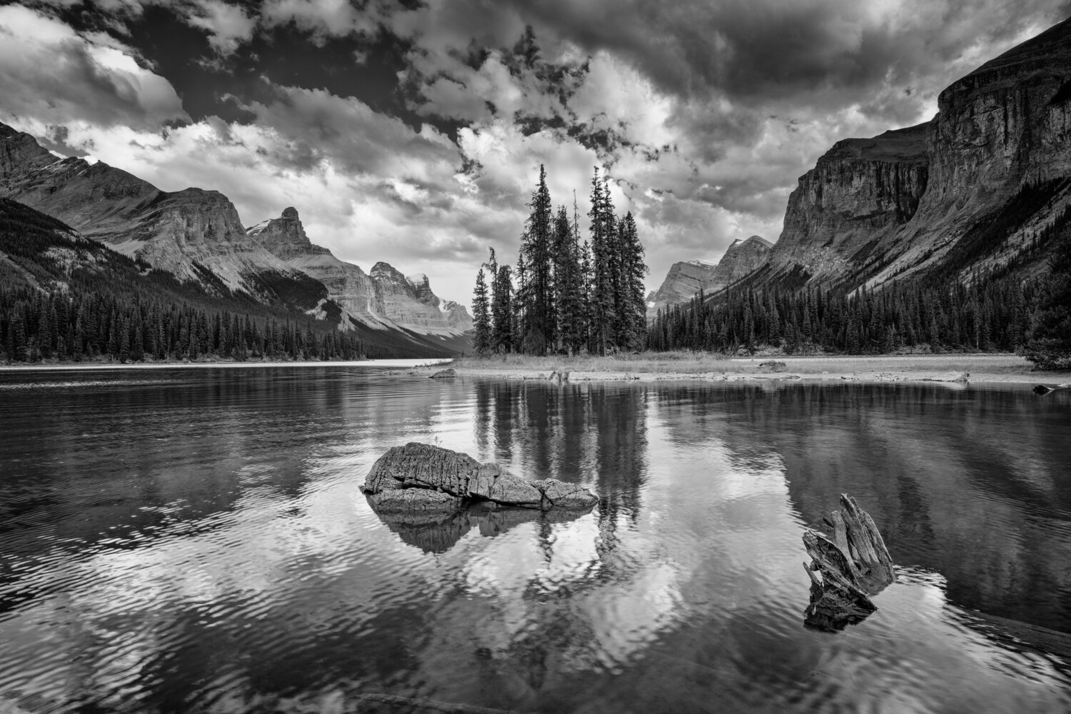 Black and white landscape of a pristine alpine lake with evergreen islands and dramatic mountain peaks