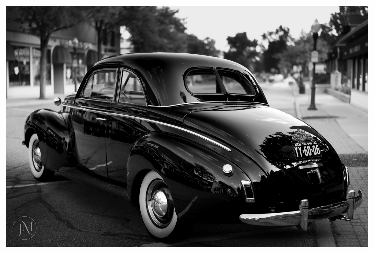 Black vintage automobile with white-wall tires parked on a tree-lined street in black and white