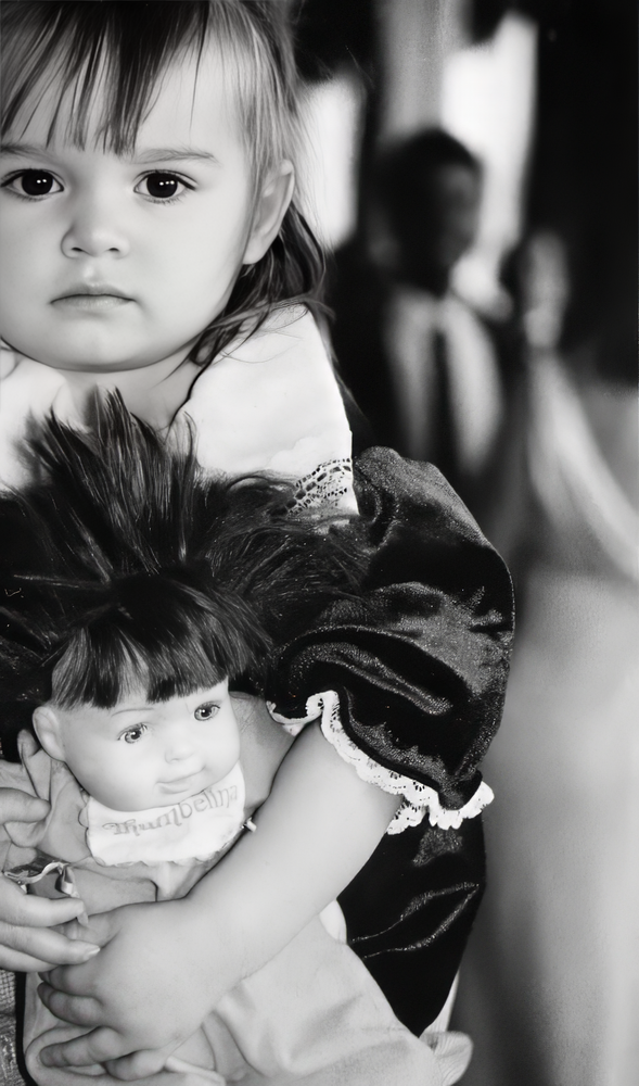 Black and white portrait of a young child with bangs holding a dark-haired doll and gazing solemnly at the camera.