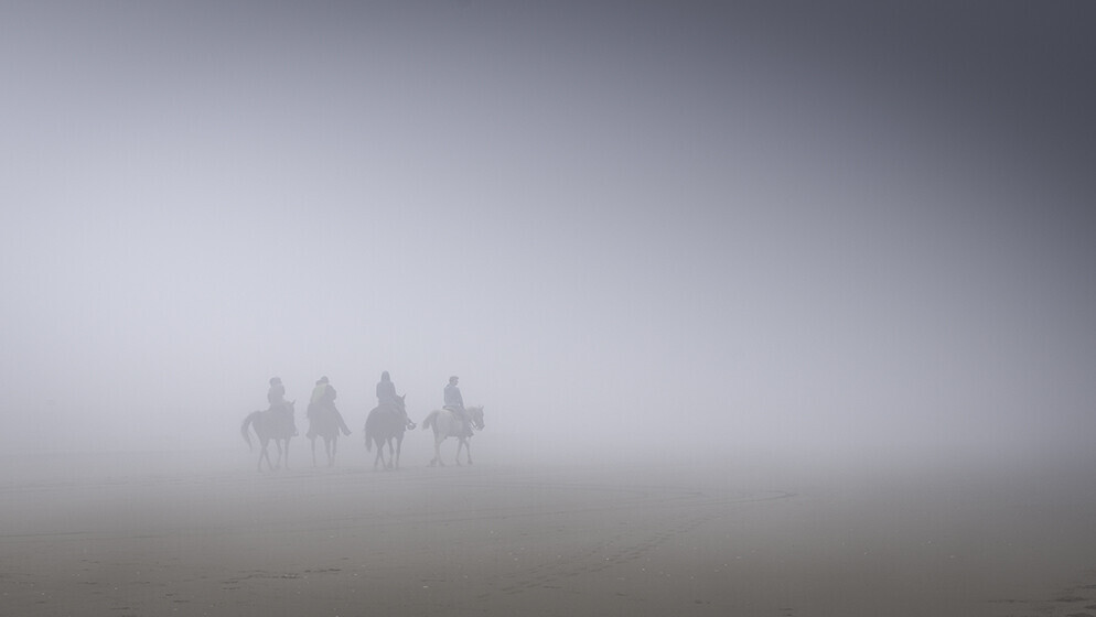 Four riders on horseback crossing a misty beach, silhouetted against a pale fog with no visible horizon.