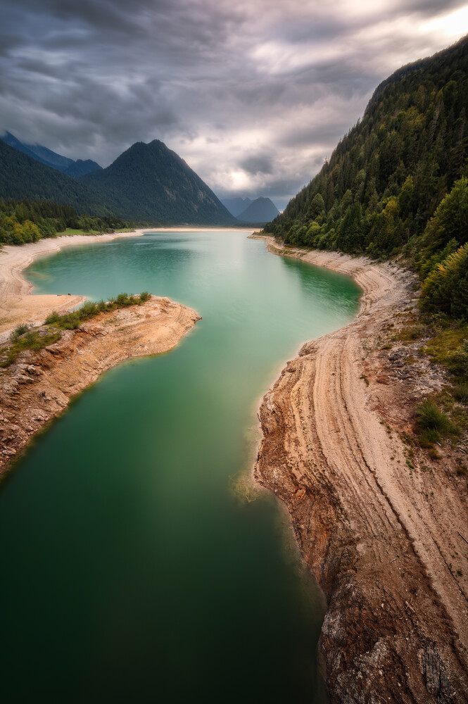 Turquoise alpine lake surrounded by forested mountains and rocky shores under dramatic clouds